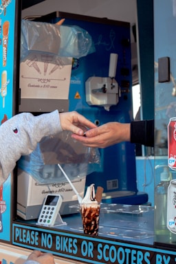 Two hands are engaged in a transaction at an ice cream truck, with one hand receiving change. The truck's interior is visible, showing an ice cream dispenser, a carton box labeled as containing cones, and various stickers on the window. A cup of ice cream with chocolate sauce and a flake is placed on the counter next to a card reader device.