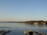 A serene lakeside scene with calm waters reflecting the clear blue sky. The shoreline is lined with trees and several houses, including a prominent large house with a light facade. Wooden docks extend into the water, with some being empty and others having small structures or posts.