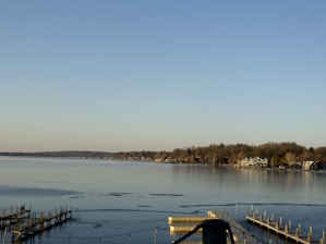 A serene lakeside scene with calm waters reflecting the clear blue sky. The shoreline is lined with trees and several houses, including a prominent large house with a light facade. Wooden docks extend into the water, with some being empty and others having small structures or posts.