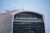 A modern skyscraper featuring a distinct curved facade with horizontal and vertical lines. The top of the building is adorned with cranes and the structure showcases extensive use of glass windows reflecting light.