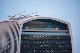 A modern skyscraper featuring a distinct curved facade with horizontal and vertical lines. The top of the building is adorned with cranes and the structure showcases extensive use of glass windows reflecting light.