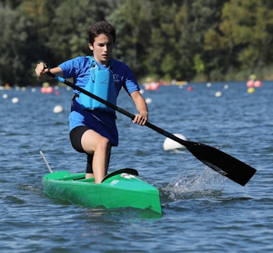 A young person paddles vigorously in a single kayak on a lake surrounded by trees. The kayak is bright green, and the individual is wearing a blue life jacket and using a black paddle. The water is slightly rippled, and there are colorful buoys scattered across the lake in the background.