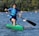 A young person paddles vigorously in a single kayak on a lake surrounded by trees. The kayak is bright green, and the individual is wearing a blue life jacket and using a black paddle. The water is slightly rippled, and there are colorful buoys scattered across the lake in the background.