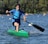 A young person paddles vigorously in a single kayak on a lake surrounded by trees. The kayak is bright green, and the individual is wearing a blue life jacket and using a black paddle. The water is slightly rippled, and there are colorful buoys scattered across the lake in the background.