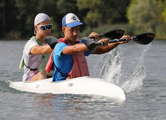 Two individuals are paddling a white kayak on a calm body of water. They are wearing athletic gear, including caps and sunglasses, suggesting engagement in a sporting activity. Water splashes are visible from the paddles, indicating motion and effort. The background features lush green foliage.