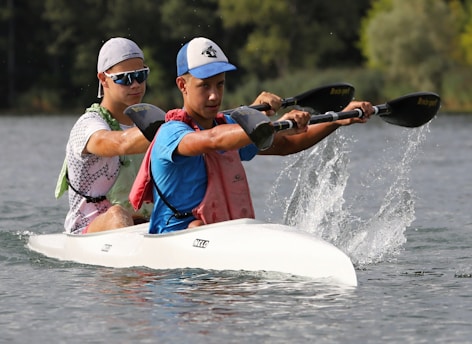 Two individuals are paddling a white kayak on a calm body of water. They are wearing athletic gear, including caps and sunglasses, suggesting engagement in a sporting activity. Water splashes are visible from the paddles, indicating motion and effort. The background features lush green foliage.