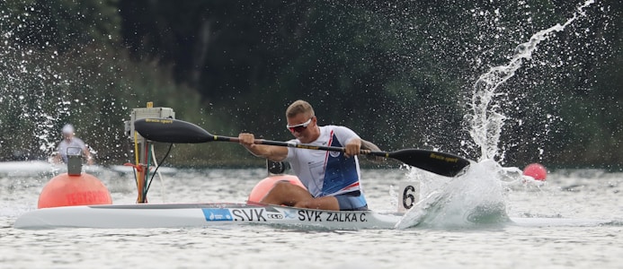 A kayaker is paddling intensely on a body of water, creating splashes. They are wearing a blue and white jersey and sunglasses. The kayak has the word 'SVK' and 'ZALKA C.' printed on it. There are buoys visible in the background on the water.