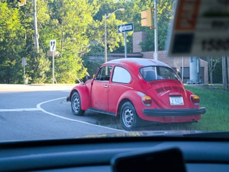 A vintage red Volkswagen Beetle is parked slightly off the road near a lush green area with trees. The car has a Virginia license plate and is situated near road signs indicating a one-way direction and a left turn. The setting appears to be a sunny day.