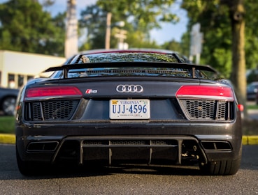 A sleek, black sports car is parked on the road. It features distinctive red taillights, a Virginia license plate, and a visible Audi R8 badge. The vehicle is surrounded by an outdoor setting with trees and a blurred background.
