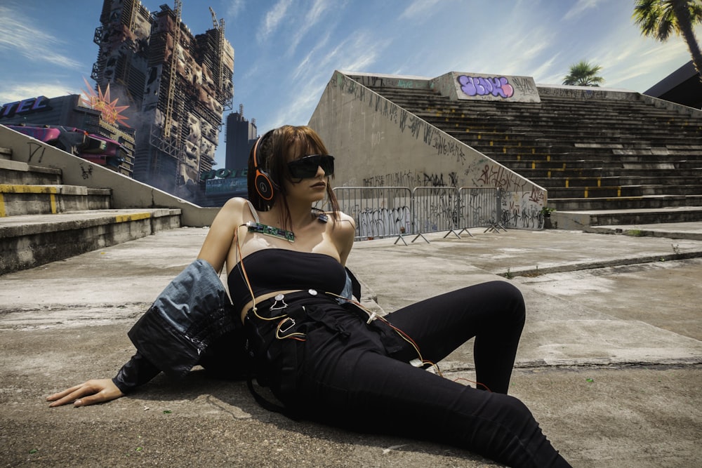 Frau in schwarzen Jeans und schwarzer Sonnenbrille sitzt tagsüber auf grauem Betonboden