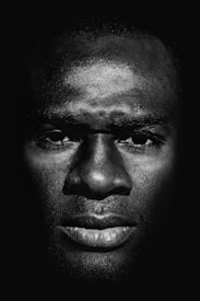 A stark and dramatic black and white portrait of a man's face with intense lighting that highlights the texture and features. The eyes and lips are especially prominent, while the rest of the background fades into darkness.