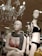 Two mannequins are displayed in a retail store setting. One wears a pink bra with a black and white patterned scarf, while the other is dressed in a black bikini and a floral-patterned jacket. Overhead, an ornate chandelier with dangling crystals is illuminated. The background shows a store environment with various clothing items visible on racks.