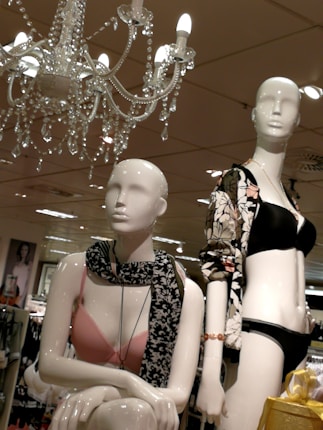 Two mannequins are displayed in a retail store setting. One wears a pink bra with a black and white patterned scarf, while the other is dressed in a black bikini and a floral-patterned jacket. Overhead, an ornate chandelier with dangling crystals is illuminated. The background shows a store environment with various clothing items visible on racks.