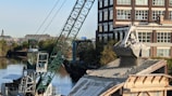 A large crane is actively unloading gravel or similar material onto a rooftop, with a building and a canal visible in the background. The crane's arm is extended over the roof, controlling a large metal scoop releasing the granular substance. Trees line the canal creating a greenscape contrast against the urban environment.