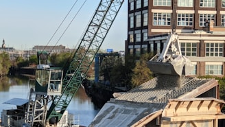 A large crane is actively unloading gravel or similar material onto a rooftop, with a building and a canal visible in the background. The crane's arm is extended over the roof, controlling a large metal scoop releasing the granular substance. Trees line the canal creating a greenscape contrast against the urban environment.
