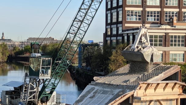 A large crane is actively unloading gravel or similar material onto a rooftop, with a building and a canal visible in the background. The crane's arm is extended over the roof, controlling a large metal scoop releasing the granular substance. Trees line the canal creating a greenscape contrast against the urban environment.
