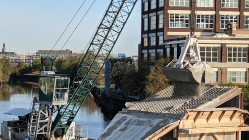 A large crane is actively unloading gravel or similar material onto a rooftop, with a building and a canal visible in the background. The crane's arm is extended over the roof, controlling a large metal scoop releasing the granular substance. Trees line the canal creating a greenscape contrast against the urban environment.