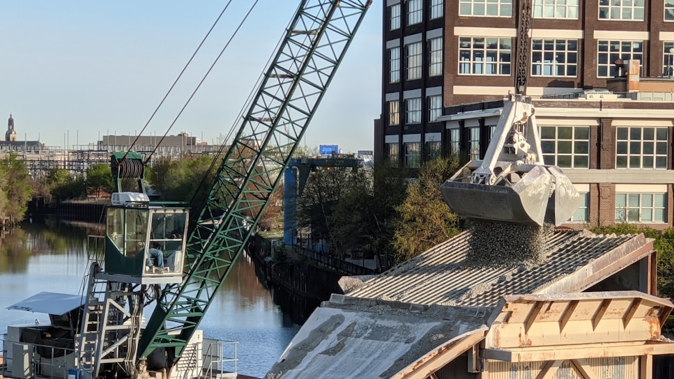 A large crane is actively unloading gravel or similar material onto a rooftop, with a building and a canal visible in the background. The crane's arm is extended over the roof, controlling a large metal scoop releasing the granular substance. Trees line the canal creating a greenscape contrast against the urban environment.