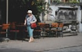 An elderly woman with a cane walks past a row of outdoor wooden chairs and tables on a cobblestone path. She is wearing a floral blouse, turquoise pants, a sun hat, and carrying a red bag. In the background, there is an old stone wall with a decorative, weathered architectural structure, partially covered with ivy.