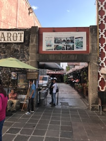 An outdoor market area featuring a pathway lined with stalls and shops. A sign for 'Cuccina Italiana' is above a stone archway leading to more seating and shopping areas. People are walking around, and a vendor is selling food from metal pots nearby. The walls are decorated with paintings and colorful signs.