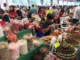A bustling market scene with various stalls displaying a colorful array of foods. Large bags and containers hold assorted snacks, including candies, dried fruits, and nuts. People are busy interacting and making purchases, with one vendor packaging goods. Bright green table coverings and colorful signs add vibrancy to the atmosphere.