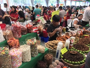 A bustling market scene with various stalls displaying a colorful array of foods. Large bags and containers hold assorted snacks, including candies, dried fruits, and nuts. People are busy interacting and making purchases, with one vendor packaging goods. Bright green table coverings and colorful signs add vibrancy to the atmosphere.