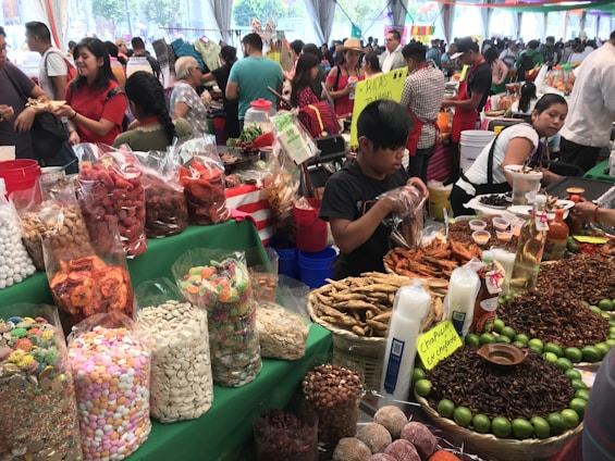 A bustling market scene with various stalls displaying a colorful array of foods. Large bags and containers hold assorted snacks, including candies, dried fruits, and nuts. People are busy interacting and making purchases, with one vendor packaging goods. Bright green table coverings and colorful signs add vibrancy to the atmosphere.