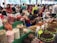 A bustling market scene with various stalls displaying a colorful array of foods. Large bags and containers hold assorted snacks, including candies, dried fruits, and nuts. People are busy interacting and making purchases, with one vendor packaging goods. Bright green table coverings and colorful signs add vibrancy to the atmosphere.