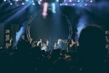 A live music performance on stage with several musicians playing instruments including drums and guitar. The stage is illuminated with colorful lights and smoke effects, creating a lively and dynamic atmosphere. The audience is visible in silhouette, indicating an engaged crowd.