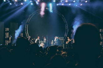 A live music performance on stage with several musicians playing instruments including drums and guitar. The stage is illuminated with colorful lights and smoke effects, creating a lively and dynamic atmosphere. The audience is visible in silhouette, indicating an engaged crowd.
