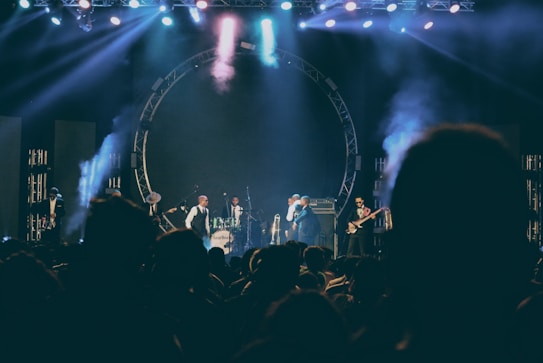 A live music performance on stage with several musicians playing instruments including drums and guitar. The stage is illuminated with colorful lights and smoke effects, creating a lively and dynamic atmosphere. The audience is visible in silhouette, indicating an engaged crowd.