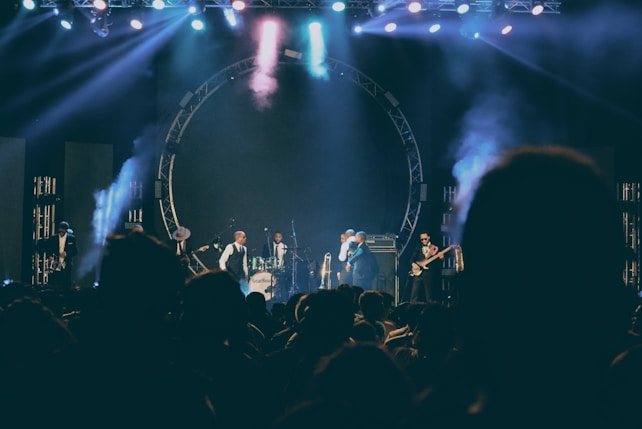 A live music performance on stage with several musicians playing instruments including drums and guitar. The stage is illuminated with colorful lights and smoke effects, creating a lively and dynamic atmosphere. The audience is visible in silhouette, indicating an engaged crowd.