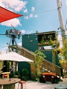 A rustic outdoor setting featuring a modern wooden structure with stairs leading up to a rooftop deck adorned with plants. An old vehicle front is repurposed as decor under the stairs, surrounded by potted greenery. The area includes wooden tables with stools and is shaded by large umbrellas. The sky is blue with scattered clouds, and a sign with the word 'CAMP' is prominently displayed on a metal frame.