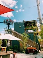 A rustic outdoor setting featuring a modern wooden structure with stairs leading up to a rooftop deck adorned with plants. An old vehicle front is repurposed as decor under the stairs, surrounded by potted greenery. The area includes wooden tables with stools and is shaded by large umbrellas. The sky is blue with scattered clouds, and a sign with the word 'CAMP' is prominently displayed on a metal frame.