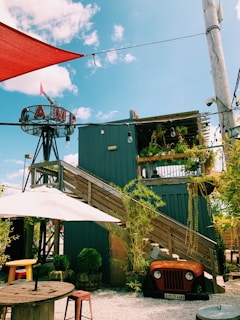 A rustic outdoor setting featuring a modern wooden structure with stairs leading up to a rooftop deck adorned with plants. An old vehicle front is repurposed as decor under the stairs, surrounded by potted greenery. The area includes wooden tables with stools and is shaded by large umbrellas. The sky is blue with scattered clouds, and a sign with the word 'CAMP' is prominently displayed on a metal frame.