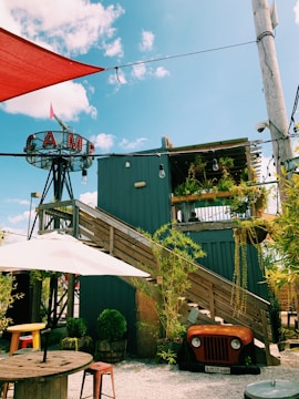 A rustic outdoor setting featuring a modern wooden structure with stairs leading up to a rooftop deck adorned with plants. An old vehicle front is repurposed as decor under the stairs, surrounded by potted greenery. The area includes wooden tables with stools and is shaded by large umbrellas. The sky is blue with scattered clouds, and a sign with the word 'CAMP' is prominently displayed on a metal frame.