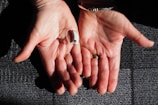 Two open hands are positioned side by side against a textured surface. A small hearing aid rests in one palm. The hands are adorned with rings and a bracelet, and the skin is illuminated by natural light, highlighting their texture.