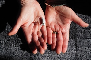 Two open hands are positioned side by side against a textured surface. A small hearing aid rests in one palm. The hands are adorned with rings and a bracelet, and the skin is illuminated by natural light, highlighting their texture.
