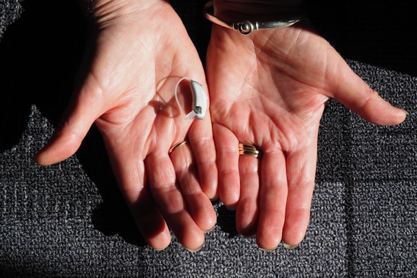 Two open hands are positioned side by side against a textured surface. A small hearing aid rests in one palm. The hands are adorned with rings and a bracelet, and the skin is illuminated by natural light, highlighting their texture.