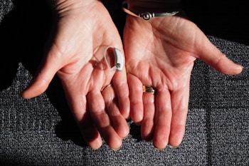 Two open hands are positioned side by side against a textured surface. A small hearing aid rests in one palm. The hands are adorned with rings and a bracelet, and the skin is illuminated by natural light, highlighting their texture.