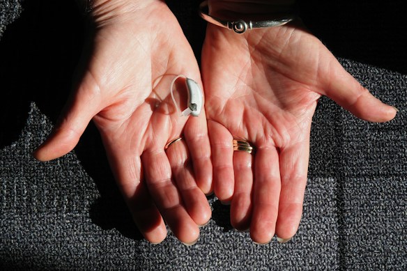 Two open hands are positioned side by side against a textured surface. A small hearing aid rests in one palm. The hands are adorned with rings and a bracelet, and the skin is illuminated by natural light, highlighting their texture.