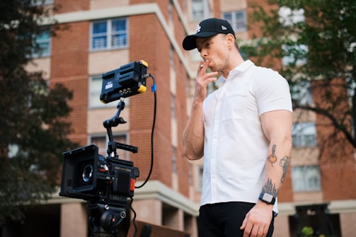 A person wearing a white shirt and black cap is smoking while standing next to a professional camera on a tripod. The background features a brown brick building with windows and some trees.