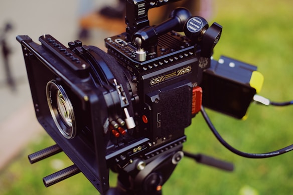 A professional-grade digital camera setup is mounted on a tripod. The camera is equipped with various accessories, including a matte box, follow focus gears, and a monitor. The focus is sharp on the camera, highlighting its detailed components and mechanical complexity. The background is blurred, featuring soft green hues, suggesting an outdoor setting.