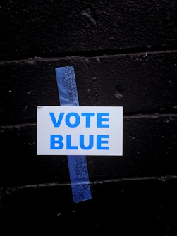 A white sign with the words 'VOTE BLUE' in blue capital letters is taped to a black brick wall using a piece of blue painter's tape.