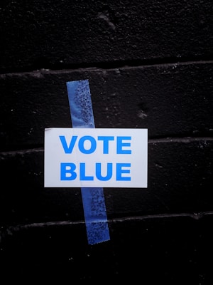 A white sign with the words 'VOTE BLUE' in blue capital letters is taped to a black brick wall using a piece of blue painter's tape.