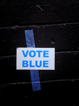 A white sign with the words 'VOTE BLUE' in blue capital letters is taped to a black brick wall using a piece of blue painter's tape.