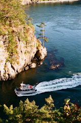 A small boat cruises through a narrow waterway surrounded by lush green trees and rocky cliffs. The clear blue water reflects the surrounding landscape, and the boat's wake creates a dynamic pattern on the water surface. The scene exudes a sense of adventure and natural beauty in a coastal environment.