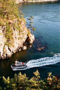 A small boat cruises through a narrow waterway surrounded by lush green trees and rocky cliffs. The clear blue water reflects the surrounding landscape, and the boat's wake creates a dynamic pattern on the water surface. The scene exudes a sense of adventure and natural beauty in a coastal environment.