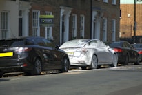 A street scene featuring several parked cars. One car is covered in white foam, suggesting it is undergoing a wash or cleaning. The background includes a brick residential building with several windows and doorways. A 'sold' sign with a phone number is visible near the building.