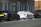 A street scene featuring several parked cars. One car is covered in white foam, suggesting it is undergoing a wash or cleaning. The background includes a brick residential building with several windows and doorways. A 'sold' sign with a phone number is visible near the building.